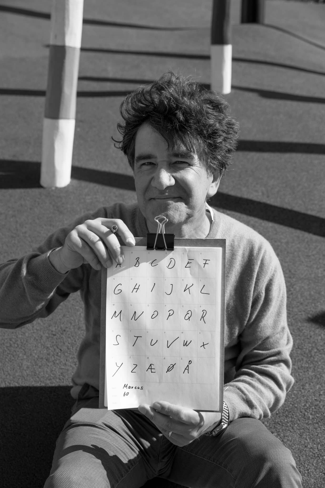 Svarthvitt foto av middelaldrende mann som holder en skriveplate med et ark hvor han har skrevet alle bokstavene i alfabetet.