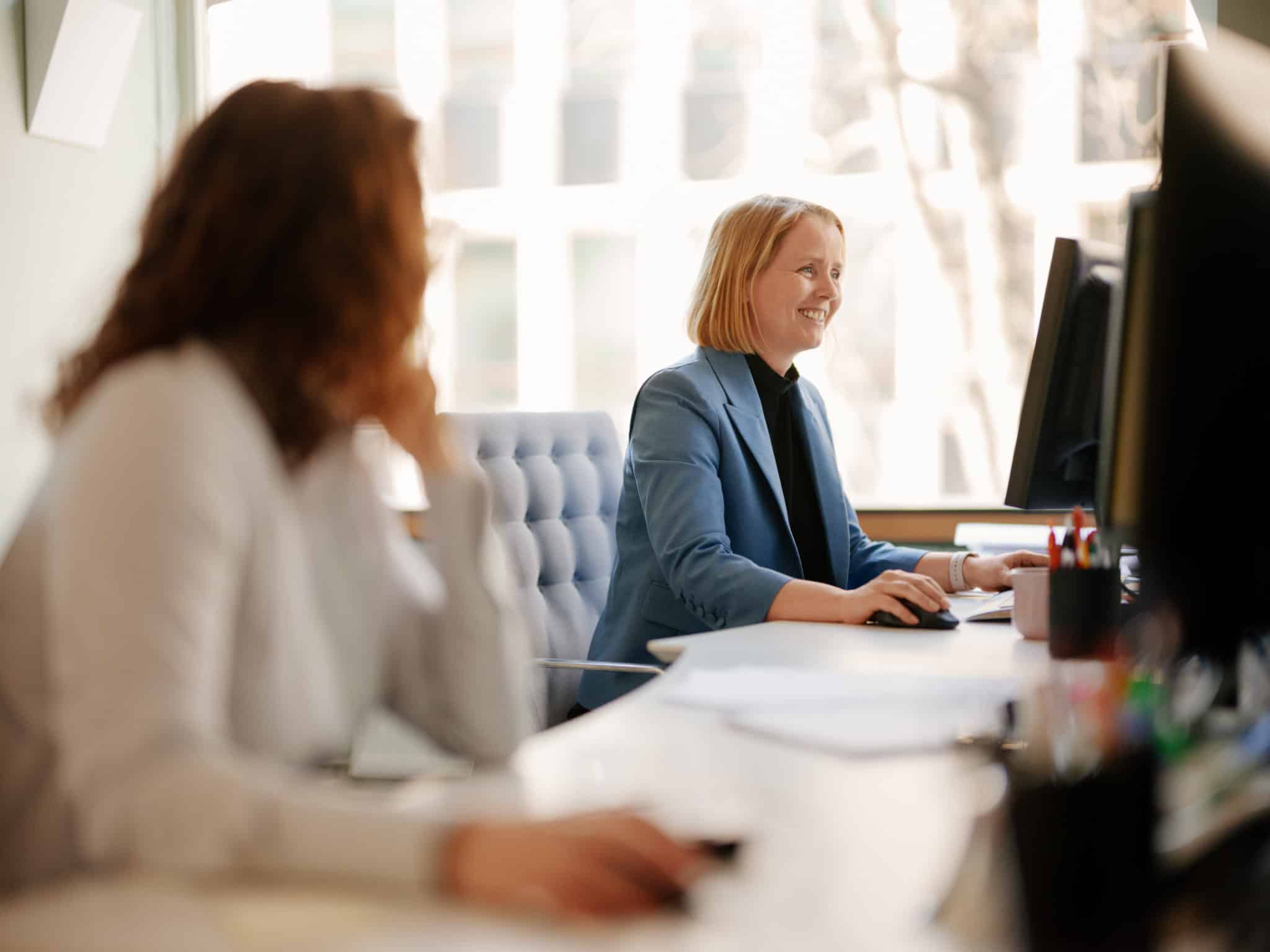 Foto av to kvinner som sitter og jobber foran hver sin dataskjerm. Kvinnen nærmest kamera er uskarp og ser mot sin kollega som sitter og smiler.