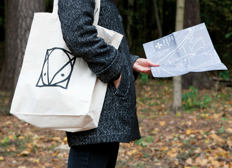 Foto av en person som står ute i en park, og med en hvit linbag med logoen til Ekebergparken. Personen holder et kart over parken i hånden.